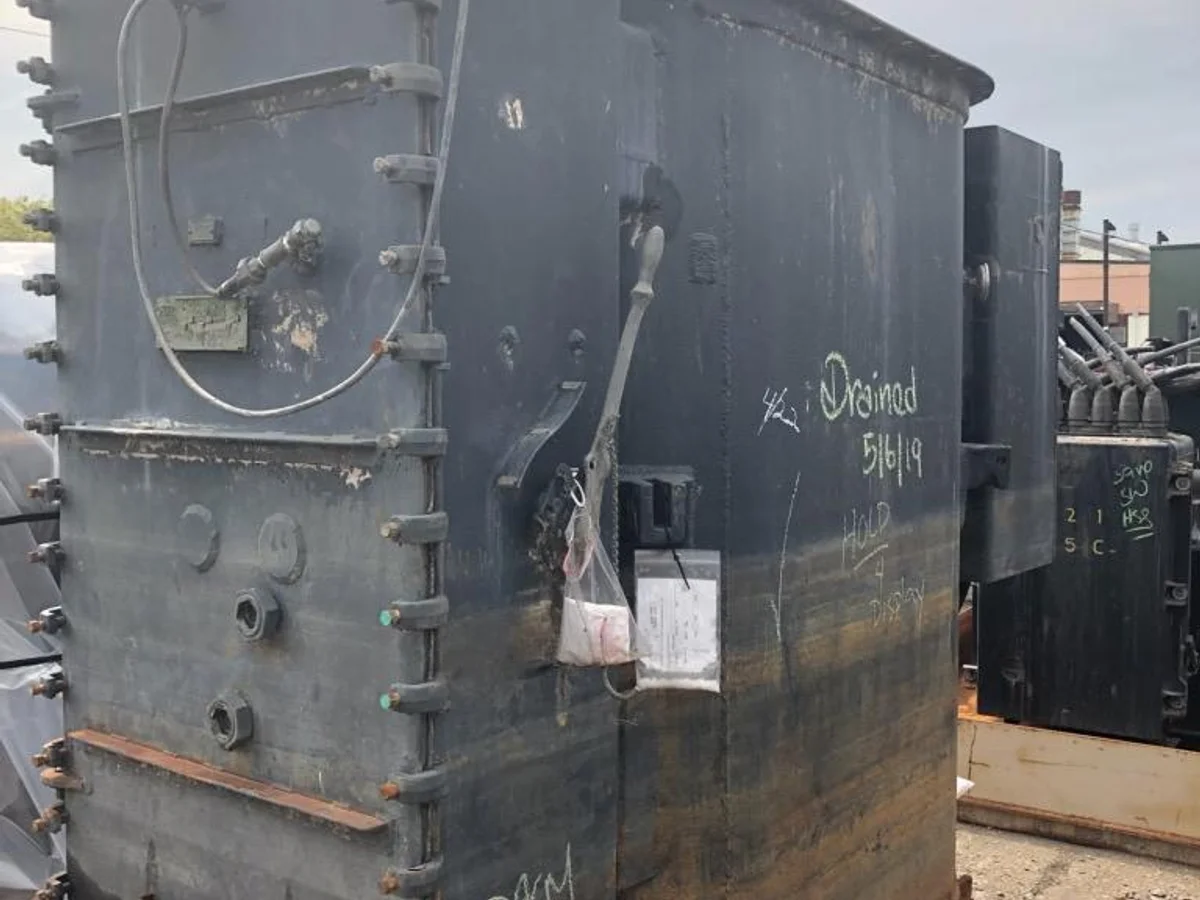 Drained, weathered transformer tank on a pallet outdoors with surface corrosion at the base