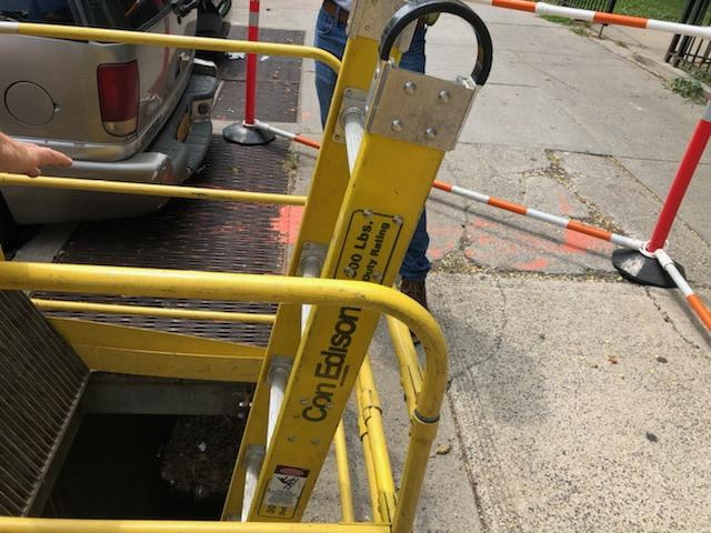 Con Edison street-level vault access — yellow safety railing, traffic cones, and barricades around an open vault